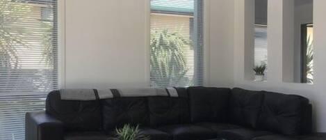 Una sala de estar moderna con un sofá de cuero negro, una mesa de centro de vidrio y ventanas grandes con persianas.
