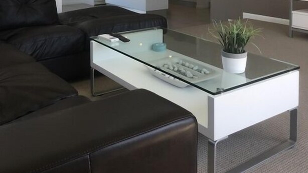 Una sala de estar moderna con una mesa de centro de vidrio, un sofá de cuero negro y una cocina al fondo.