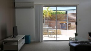 Un moderno salón con televisor de pantalla plana, sofá, mesa de centro y puerta corrediza de vidrio que da a un patio exterior con mesa y sillas.