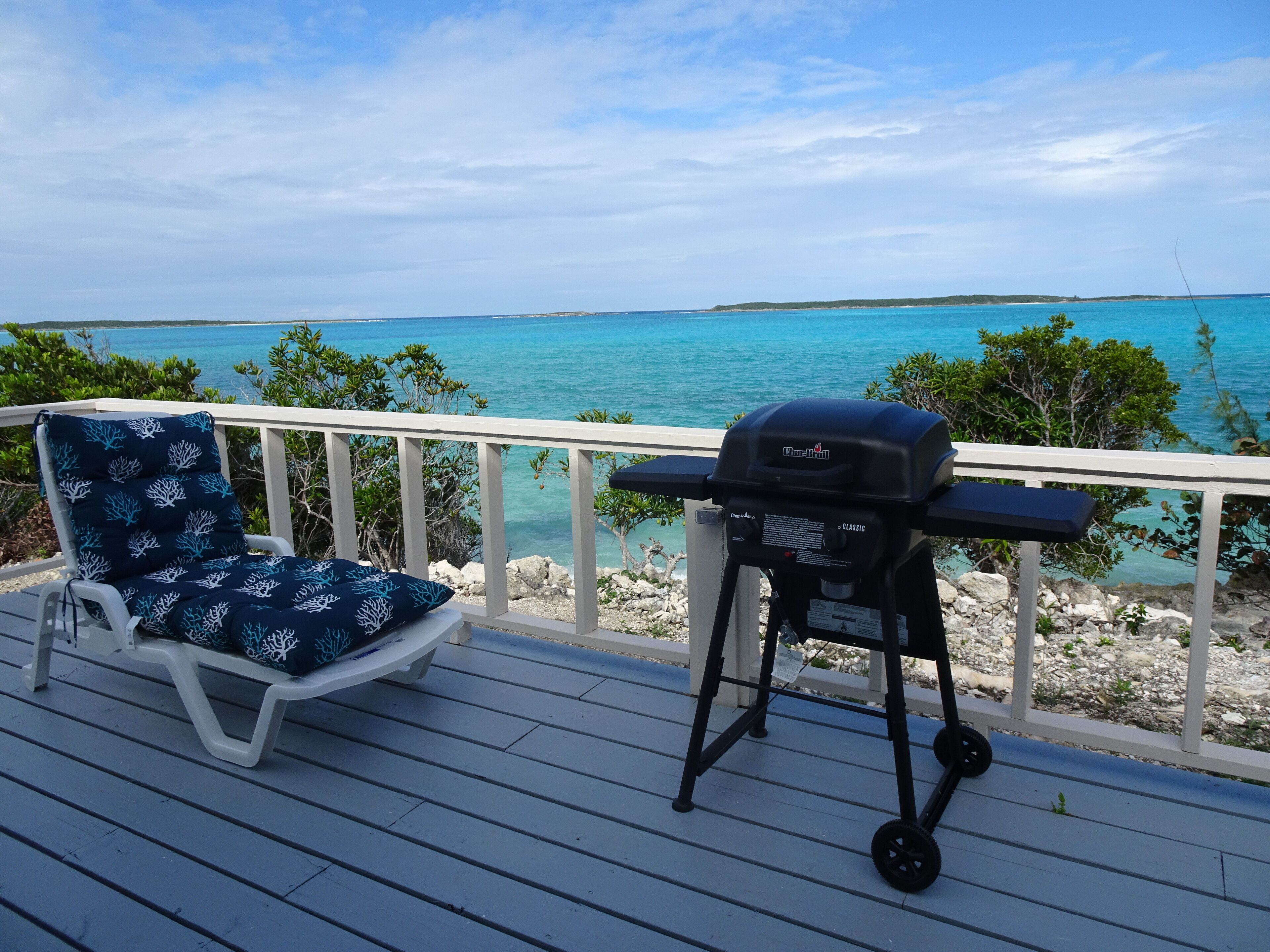 A deck with a grill and a lounge chair, overlooking a clear blue sea.