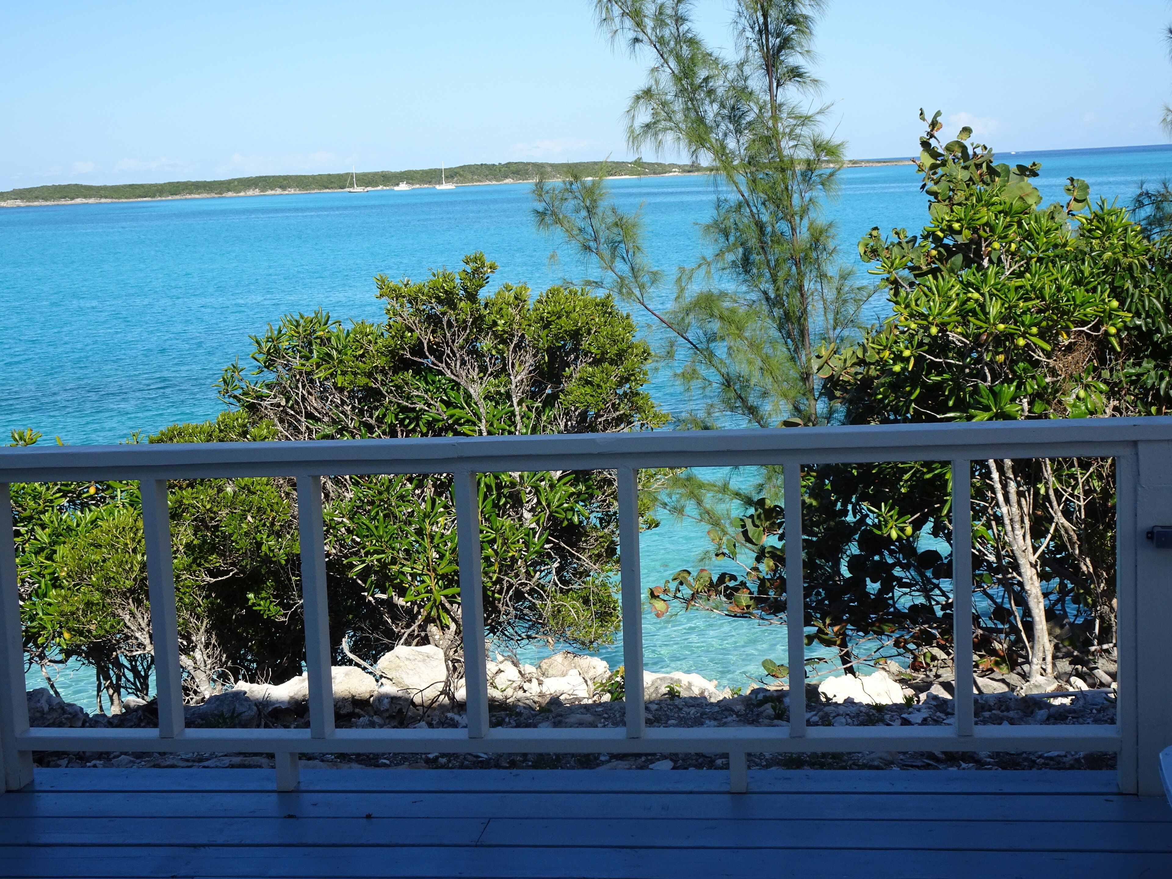 A view from a balcony with a railing, overlooking a clear blue sea and a distant shoreline with sparse vegetation.