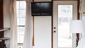 A room with a television mounted on the wall, a door with a window, a lamp, and a small table with a chair.