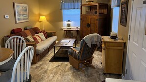 A cozy living room with a sofa, chairs, a coffee table, a wooden cabinet, a ceiling fan, and a window with blinds.