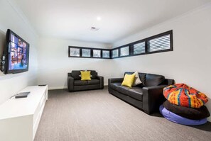 A modern living room with a flat-screen TV, two black sofas with yellow and patterned cushions, a white console table, and a large window with blinds. - Lilywood—Minutes from Wineries & Yallingup Beach (Yallingup)