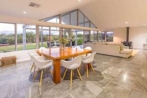 A spacious dining area with a large wooden table and white chairs, a modern kitchen with a stove, and a large window offering a view of the outdoors.
