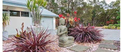 A serene garden with a Buddha statue, red foliage plants, and a building in the background.