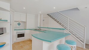 Une cuisine compacte avec des meubles blancs, un plan de travail de couleur turquoise, un four encastré et un escalier doté d’une rampe blanche.