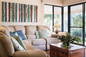 A living room with a beige sectional sofa, a wooden coffee table, and a large window with a view of greenery. - Vaila—Bushland Charm near Beaches & Top Wineries (Yallingup)