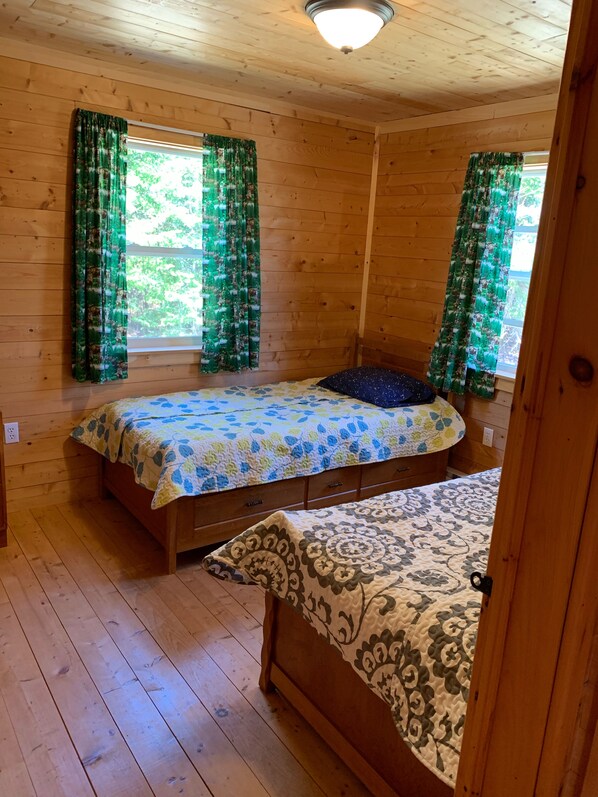 A wooden cabin room with two beds, a window with green curtains, and a wooden ceiling. - Lakefront cottage on a Wallaback lake (New Ross)