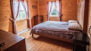 Une chambre dans une cabane en bois, avec un lit, deux fenêtres avec des rideaux, un ventilateur de plafond et une commode en bois.