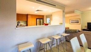 A modern kitchen with wooden cabinets, a stainless steel refrigerator, and a red bowl on the counter. There are three white bar stools and a sofa in the adjacent living area. - Surfside 9: In the Idyllic Yallingup Beach Scene (Yallingup)