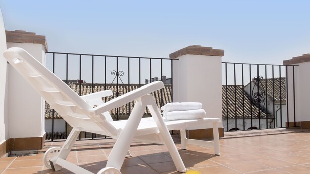 Una terraza en la azotea con dos sillones blancos, una mesita con un vaso de jugo de naranja y piso de baldosas.