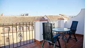 A balcony with a round glass table and two chairs, overlooking a cityscape with terracotta roofs.