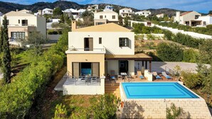 A modern villa with a swimming pool, outdoor seating, and a view of the surrounding hillside community. - Aegean Blue Villas - Aphrodite Villa (Kalathos)