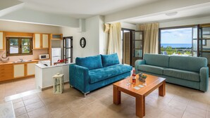 A modern living room with two sofas, a wooden coffee table, a kitchen with a microwave, and a view of the sea. - Aegean Blue Villas - Aphrodite Villa (Kalathos)