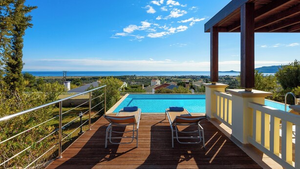 A wooden deck with two lounge chairs overlooking a swimming pool and a scenic view of the sea and distant landscape. - Aegean Blue Villas - Aphrodite Villa (Kalathos)