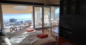 Ein Schlafzimmer mit großem Fenster, einem Bett, einem Fernseher und einem Balkon mit Stühlen.