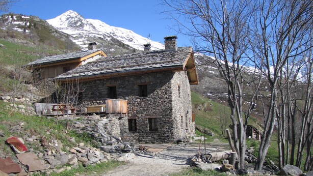 Ein Steinhaus mit einer Holzdielen-Terrasse, einem Kiesweg, der zum Eingang führt, und einer Bergkulisse im Hintergrund.