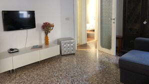 Ein Wohnzimmer mit einem Flachbildfernseher, einem weißen Konsolentisch, einem grauen Hocker, einer Blumenvase und einem dunklen Holzschrank.