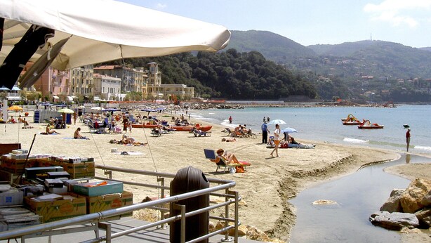 Una spiaggia con persone che prendono il sole e nuotano, una barca ormeggiata vicino alla riva e edifici sullo sfondo.