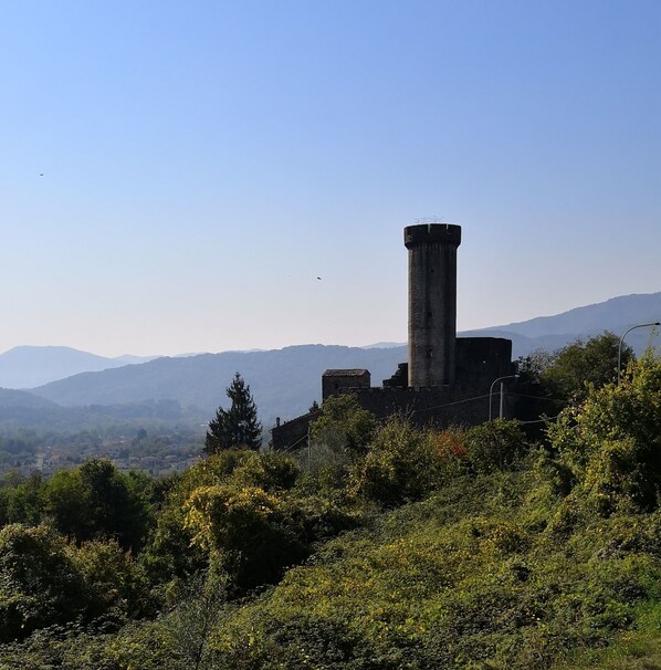 A historic tower with a cylindrical base and a flat top, surrounded by lush greenery and mountains in the background.