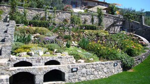 A stone building with a bell tower, surrounded by a landscaped garden with various flowers and shrubs. - Historic village house with private pool, gardens & outdoor dining, near Bagnone (Bagnone Massa Carrara)