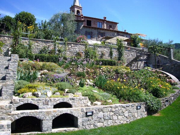 Un bâtiment en pierre doté d’un clocher, entouré d’un jardin aménagé où l’on trouve différentes espèces de fleurs et d’arbustes.