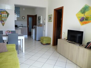 A modern living room with a green sofa, a TV, and a wooden cabinet. There is a dining area with a table and chairs, and a kitchen in the background.