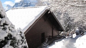 Una cabaña de madera cubierta de nieve, con techo inclinado, rodeada de árboles y montañas al fondo.