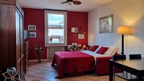 A bedroom with a bed, a desk, a chair, a ceiling fan, and a window with a view of buildings.