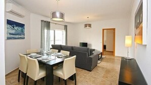 Ein moderner Essbereich mit einem Tisch für vier Personen, einem grauen Sofa und einem Fernseher im Hintergrund.
