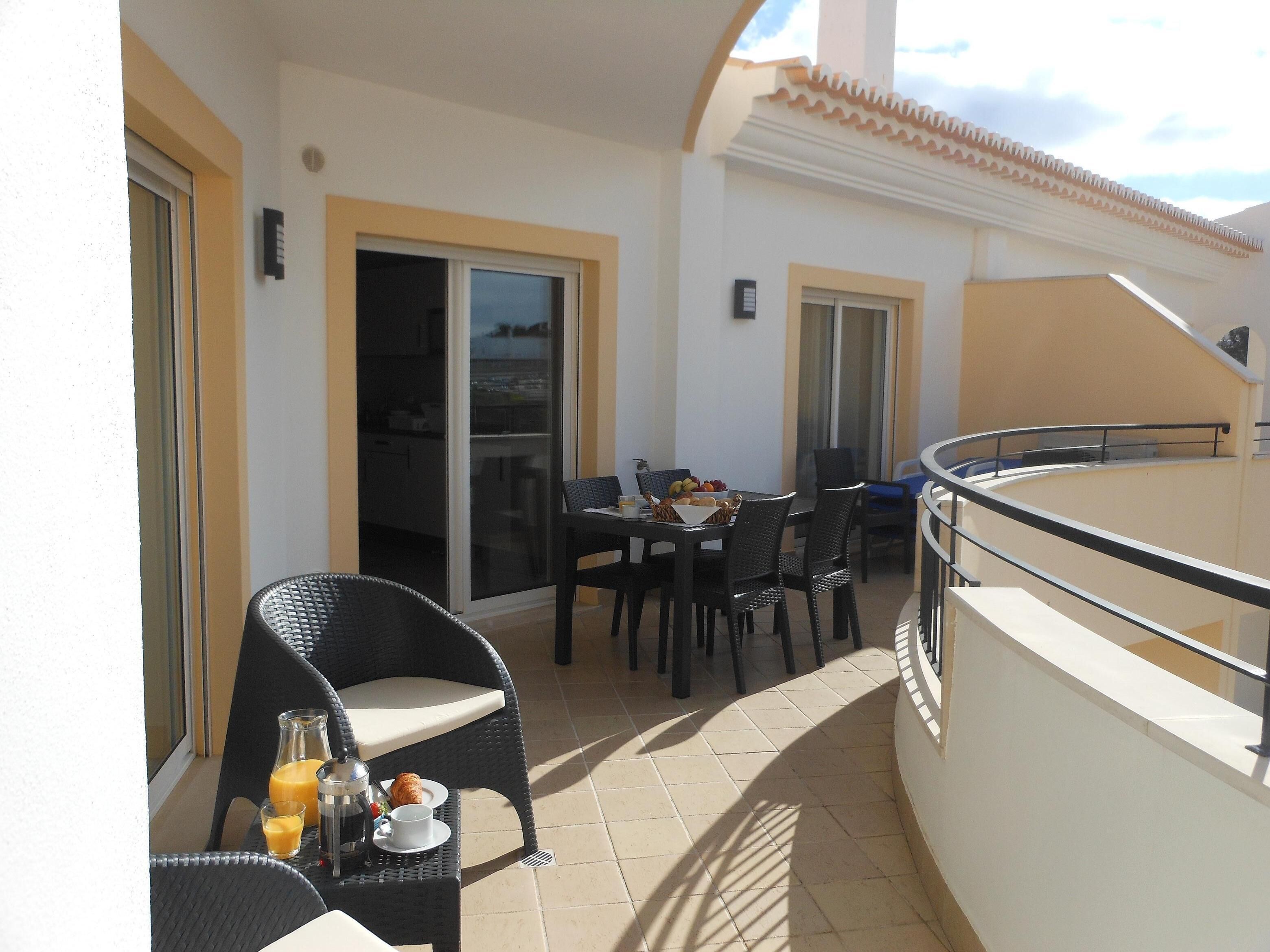 A balcony with a dining table set for two, chairs, and a small table with a drink and pastry.