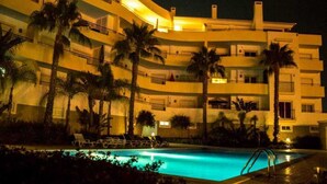Ein mehrstöckiges Hotel mit Swimmingpool, Palmen und Liegestühlen.