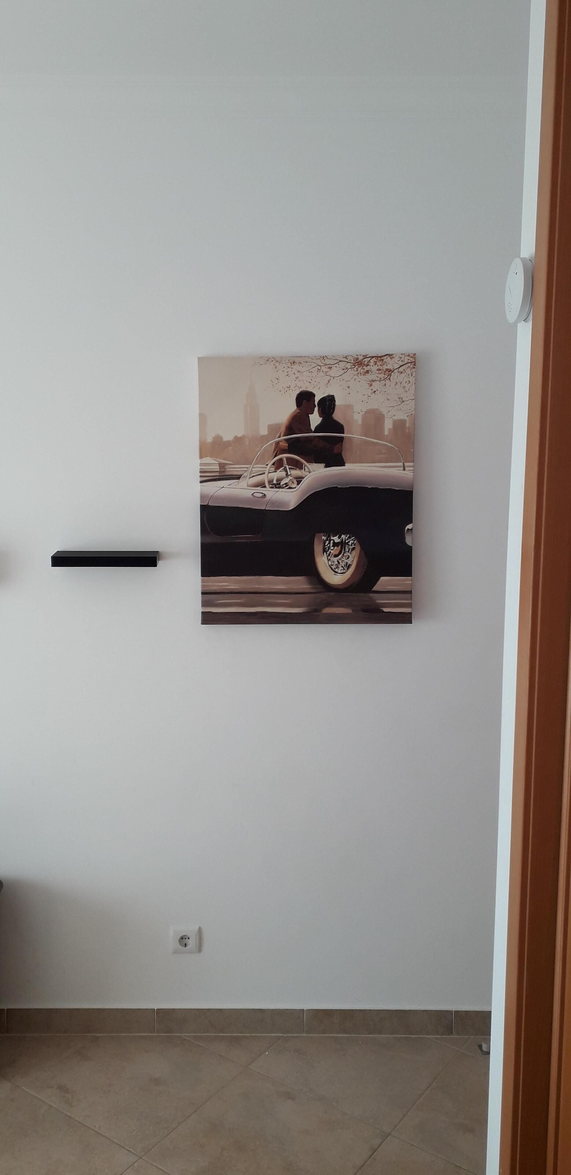 Une pièce dans laquelle on trouve une œuvre d’art représentant une voiture ancienne, accrochée au mur, ainsi que deux personnes, une prise de courant et une porte.