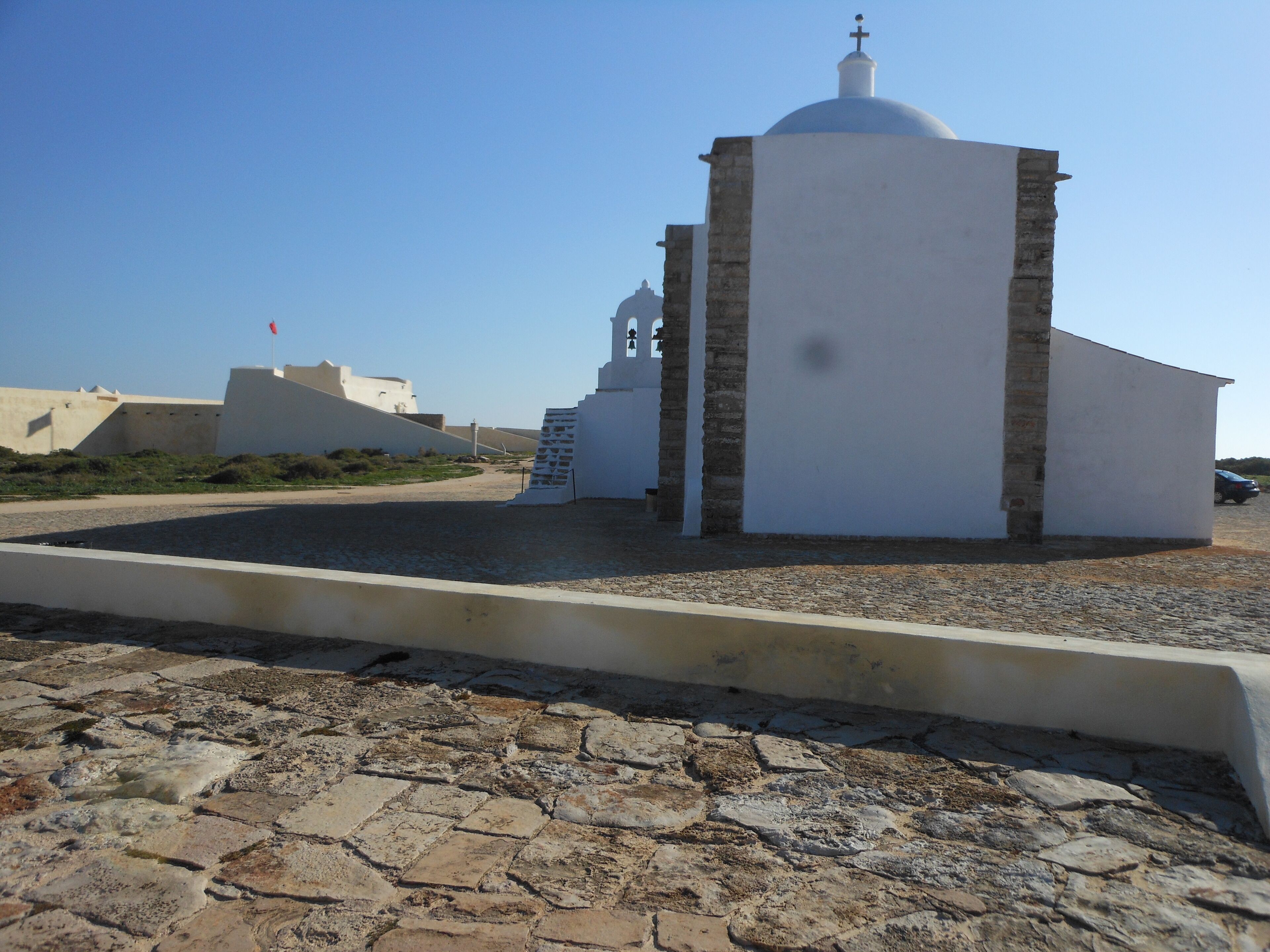 A white building with a dome and a bell tower, surrounded by a gravel area and other structures.