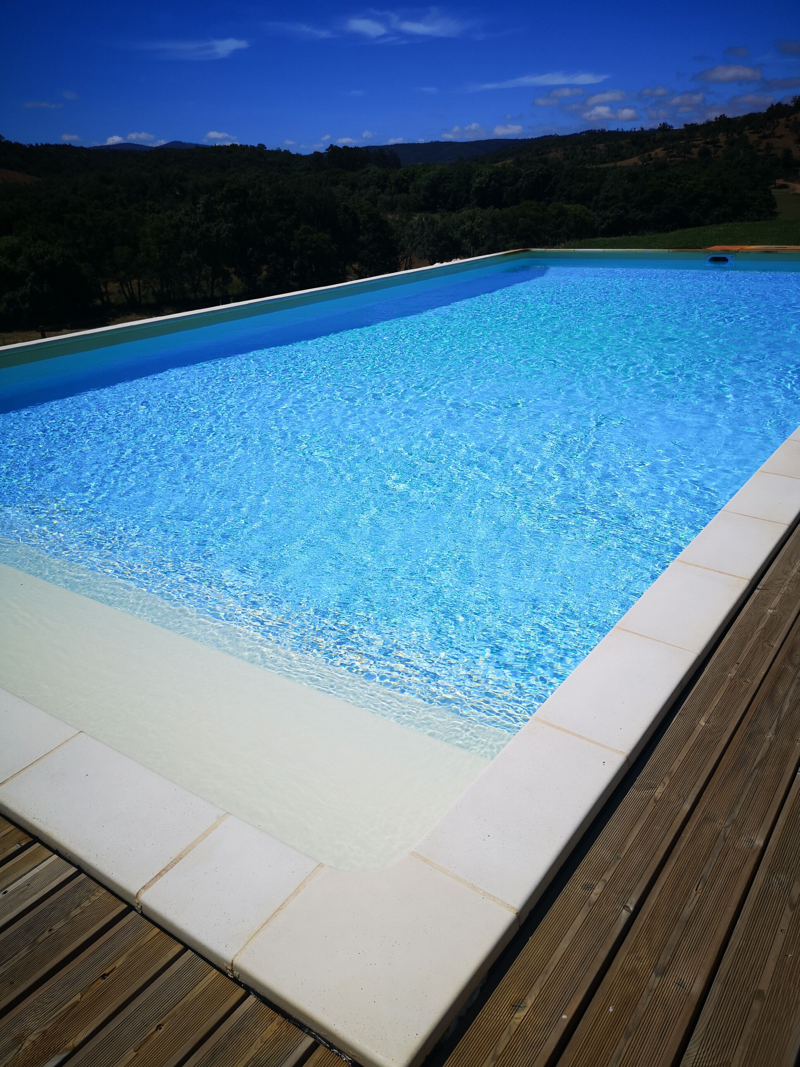 A rectangular swimming pool with clear blue water, surrounded by a wooden deck and a white tiled border.