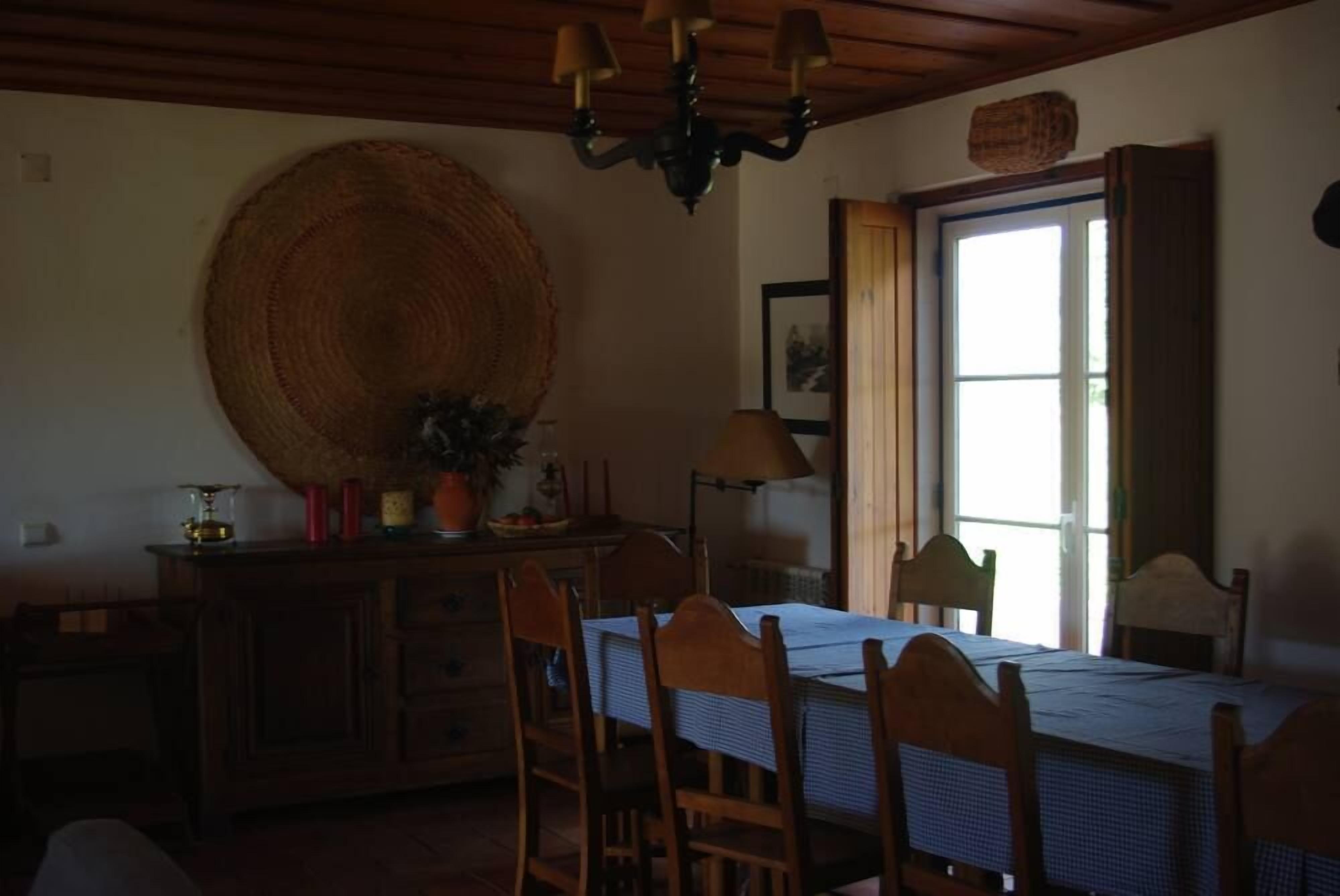 Comedor con mesa dispuesta para cuatro, sillas de madera, candelabro, una cesta grande de mimbre y una ventana con cortinas.
