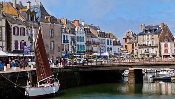 Escena frente al agua con un velero amarrado cerca de un puente, edificios de diversos estilos arquitectónicos y una calle concurrida con peatones y vehículos.