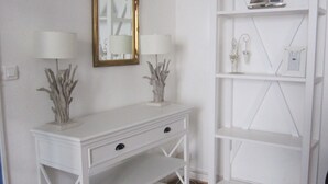 Una consola blanca con dos lámparas y un espejo, adyacente a un estante blanco con objetos decorativos.
