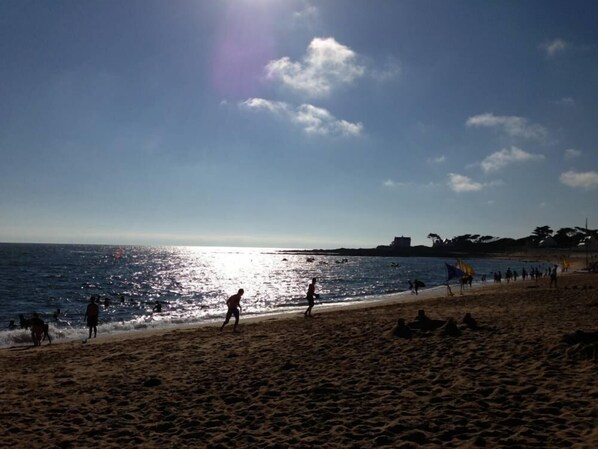 A beach with people walking along the shore, a clear sky, and the sun reflecting off the water.