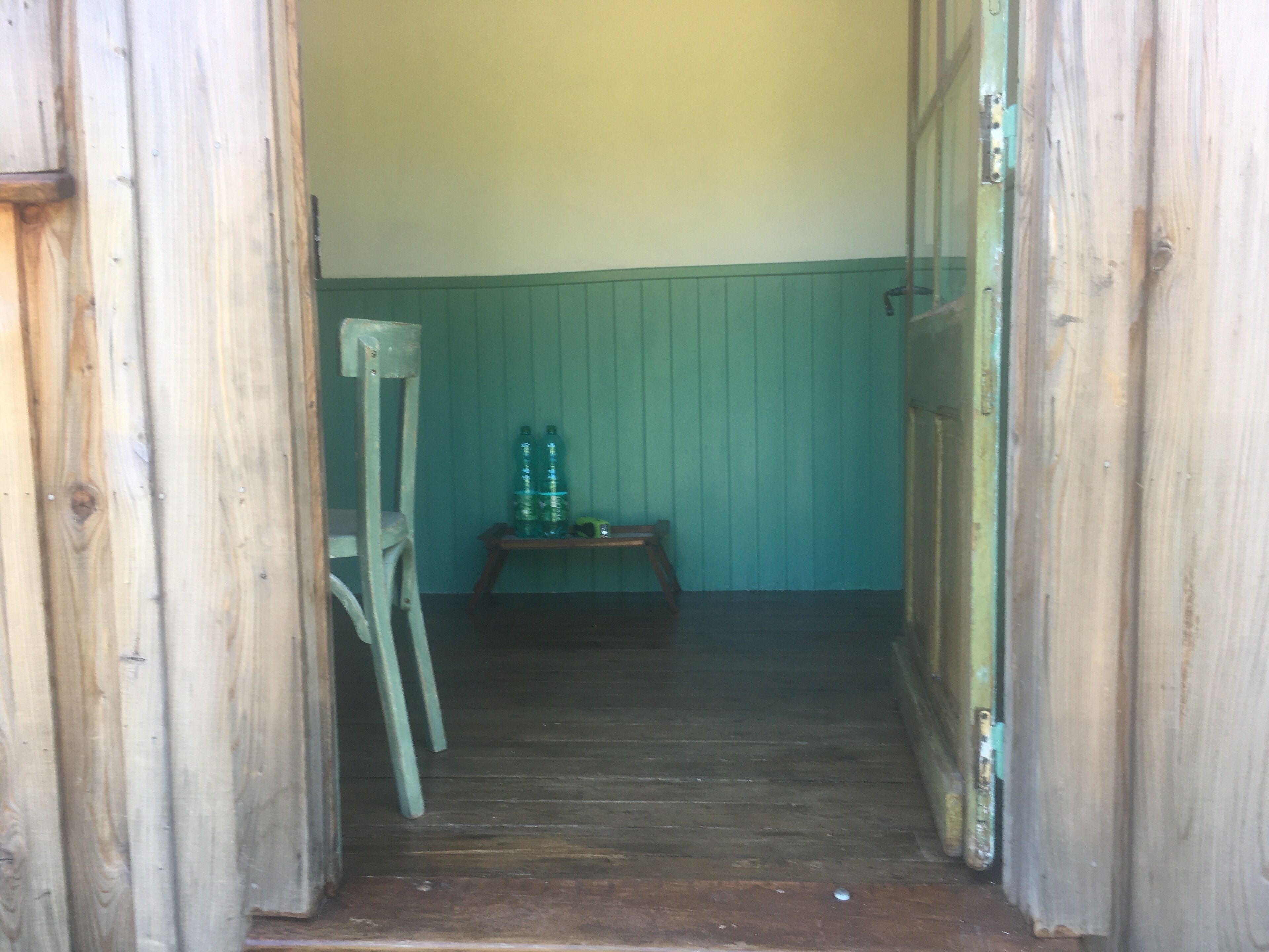 Un couloir étroit en bois, avec un mur vert et une porte en bois sur la droite. On aperçoit au fond une chaise en bois et une petite table sur laquelle sont posées deux bouteilles vertes.