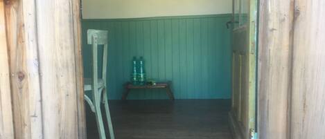 A narrow wooden hallway with a green wall and a wooden door on the right. A wooden chair and a small table with two green bottles are visible in the background.
