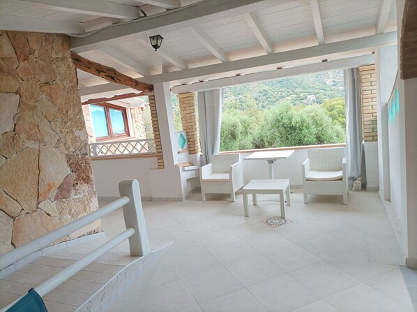 A spacious patio with white wicker furniture, a stone wall, and a view of greenery.