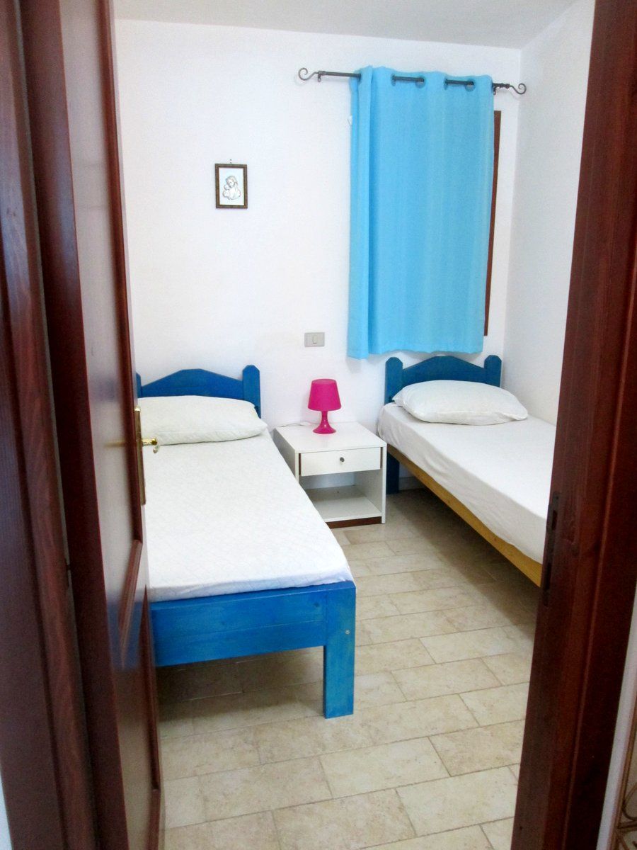 Una habitación pequeña y limpia con dos camas individuales, una mesita de noche, una lámpara y una ventana con cortina.