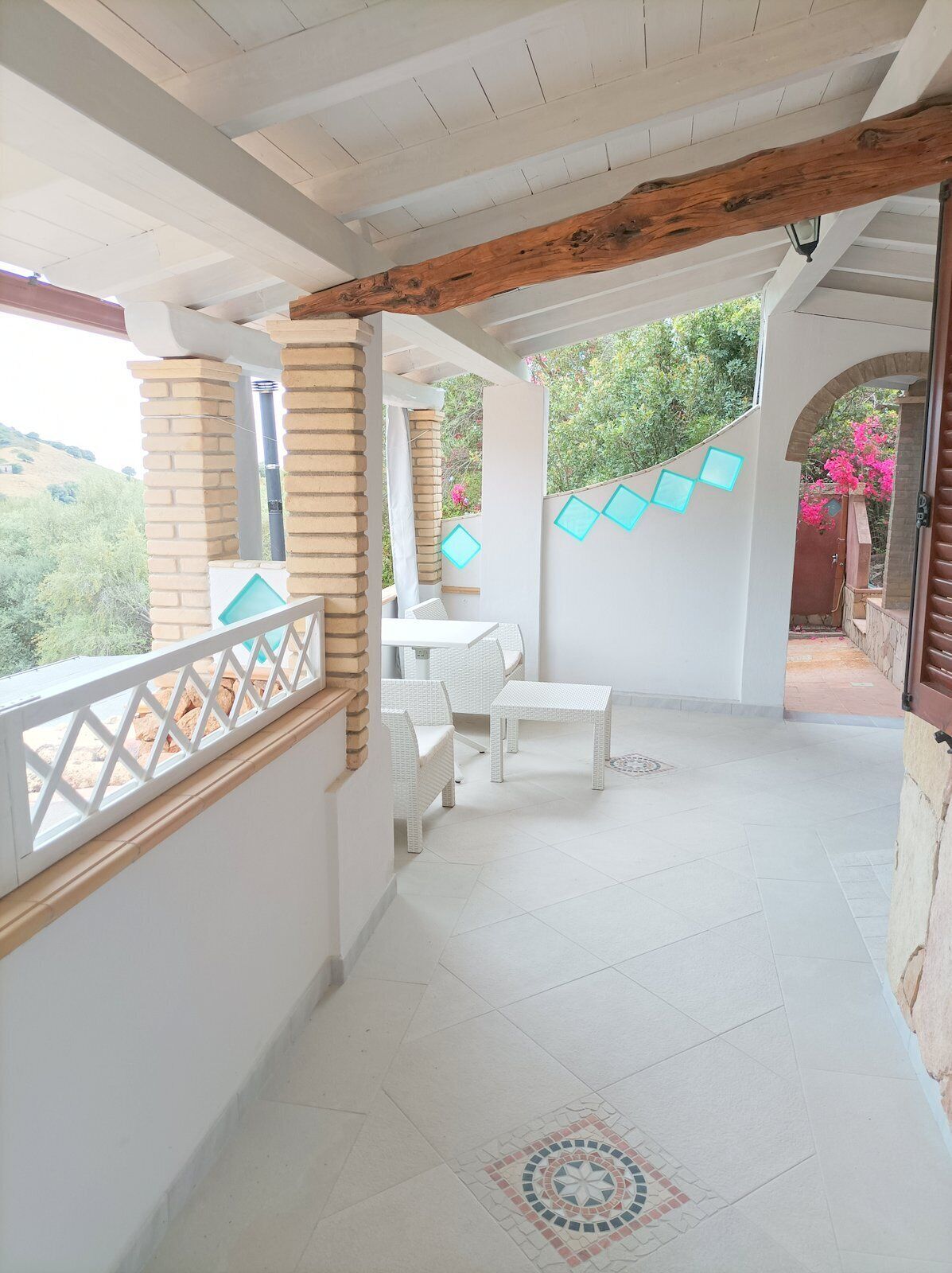 A covered balcony with white wicker furniture, a tiled floor with a decorative pattern, and a view of greenery.