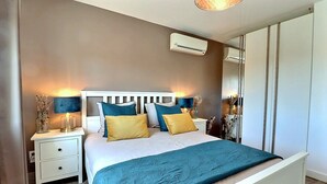 Ein Schlafzimmer mit einem weißen Bett, Kissen in Türkis und Gelb, einem weißen Nachttisch mit Lampe und einem Kleiderschrank.