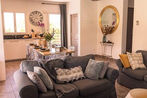 En stue med en grå sofa, et spisebord med stole, et køkken i baggrunden og et stort spejl på væggen.