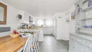 A modern kitchen with white cabinetry, a wooden dining table, and a central island. - Breakwater Cottage - Five Bedroom Cottage, Sleeps 10 (Bude)