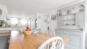 A spacious kitchen with white cabinetry, a wooden dining table, and a bowl of fruit. - Breakwater Cottage - Five Bedroom Cottage, Sleeps 10 (Bude)