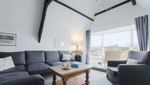 A modern living room with a grey sofa set, a wooden coffee table, and a view of the beach through large windows. - Breakwater Cottage - Five Bedroom Cottage, Sleeps 10 (Bude)